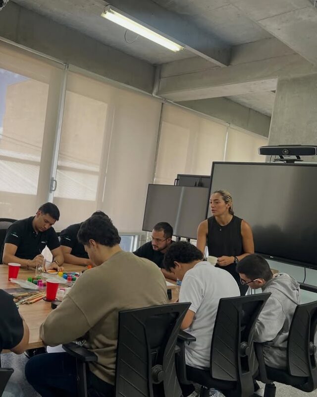 DebAnanda facilita un taller de mandalas en una sala de juntas, junto a la pantalla, con participantes alrededor de la mesa.
